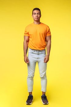 Full-length vertical shot masculine hipster guy, hispanic ethnicity, standing casually yellow background, wear stylish orange t-shirt, white pants, look camera with no expression, slightly smiling.