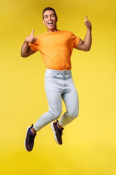 Full-length vertical shot cheerful masculine hipster guy in trendy orange t-shirt, white pants, jumping from happiness and joy, show thumbs-up gesture smiling in approval, celebrating awesome news.