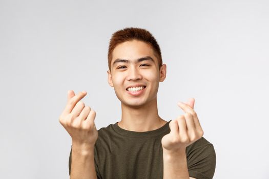 People, different expressions and lifestyle concept. Cute asian guy with stylish haircut, showing korean finger hearts and smiling lovely, express sympathy or like, stand grey background.