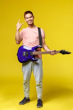 Lifestyle, leisure and youth concept. Vertical shot of enthusiastic teenage guy having fun playing electric guitar, show rock-n-roll sign and smiling, stand yellow background, play in band.