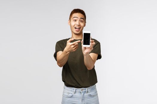 Technology, online lifestyle and communication concept. Outgoing friendly asian man showing mobile phone application, pointing at smartphone screen and look camera amused, grey background.