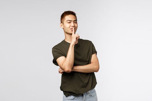 Portrait of cheeky handsome asian boyfriend prepared surprised, wink sassy and assured, smiling as shushing, promise keep secret, ask be quiet and not talk about it, grey background.