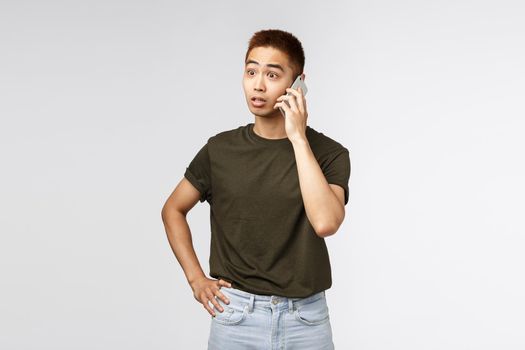 Technology, online lifestyle and communication concept. Shocked and speechless asian guy talking on phone, react startled from awesome news, having conversation via smartphone, grey background.