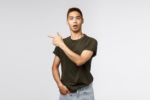 Portrait of surprised, curious asian male staring camera startled, asking question and pointing finger left at interesting strange item in store, showing way, standing grey background.