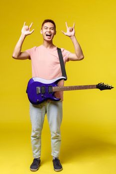 Lifestyle, leisure and youth concept. Vertical portrait of asian man in stylish outfit, perfoming, holding blue electric guitar, show rock-n-roll sign, shouting pleased, feel excitement part of band.