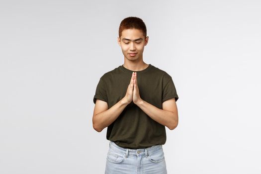 Portrait of hopeful young asian guy having faith, close eyes and smiling, clasp hands together in pray, pleading to god, want dream come true, supplicating, standing grey background.