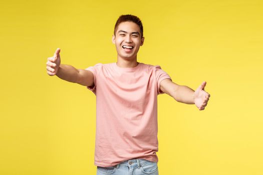 Lifestyle, travel and people concept. Portrait of cheerful, friendly asian male inviting for hug, reaching hands for embraces, stretch arms and smiling broadly, cuddling, standing yellow background.