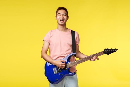 Lifestyle, leisure and youth concept. Happy asian male student playing in rock band, holding electric guitar and singing carefree with pleased smile, like perform on stage, yellow background.