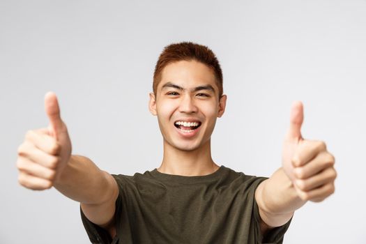 People, different expressions and lifestyle concept. Enthusiastic excited happy man, asian guy show thumbs-up and smiling, saying yes to awesome idea, approve and like concept, grey background.