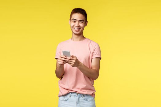 Technology, communication and lifestyle concept. Portrait of cheerful, enthusiastic hipster guy, asian male in pink t-shirt using mobile phone, laughing at camera, staying online, order delivery.