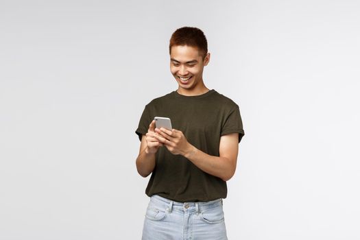 Technology, online lifestyle and communication concept. Portrait of good-looking asian young guy in green t-shirt, cheerful looking at mobile phone, texting, smiling at smartphone screen.
