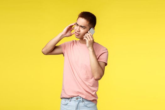 Technology, communication and lifestyle concept. Troubled asian man in pink t-shirt, feel frustrated, talking on mobile phone, squinting and grimacing bothered, stand indecisive yellow background.