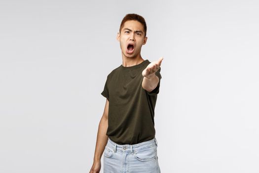 Portrait of angry complaining asian man pointing hand at camera with aggressive distressed look, disappointed arguing, received wrong parcel, being outraged standing grey background.