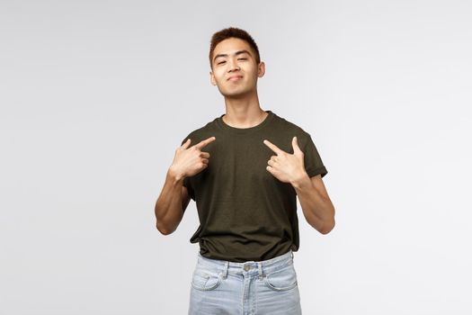 Portrait of proud and boastful young asian macho man, show-off his big ego, pointing at himself with pleased bragging smile, look camera, talking about personal achievement, grey background.