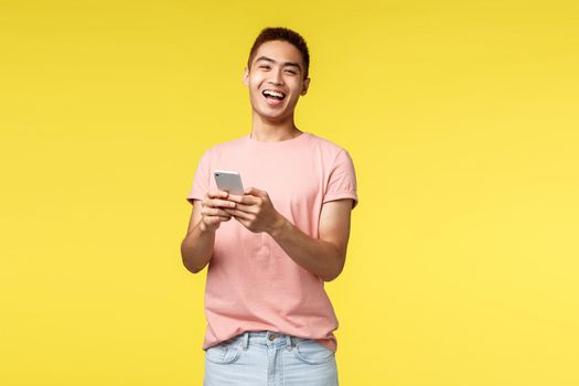 Technology, communication and lifestyle concept. Enthusiastic asian male in pink t-shirt laughing from cool joke or video, using mobile phone, smiling pleased camera, stand yellow background.
