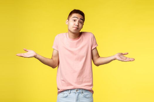 Portrait of indecisive, shrugging young asian man, spread hands sideways, smile and apologize cant help, dont know, have no plan and standing clueless yellow background. Copy space