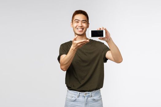 Technology, online lifestyle and communication concept. Happy proud and cheerful asian man introduce something on mobile phone, showing application, pointing smartphone display and smiling pleased.