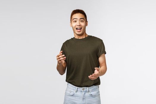 Portrait of surprised happy asian man, laughing and gasping amused, react to unexpected awesome news, express rejoice and happiness, standing grey background astonished. Copy space