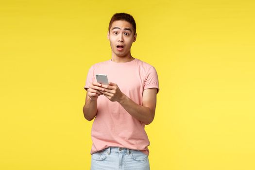 Technology, communication and lifestyle concept. Surprised asian male in pink t-shirt, holding mobile phone, open mouth gasping wondered, react to interesting news online, yellow background.