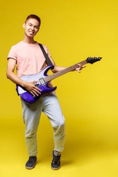 Lifestyle, leisure and youth concept. Vertical portrait of sassy handsome asian man playing electric guitar, smiling pleased, perfoming in band, enjoying jamming, yellow background.