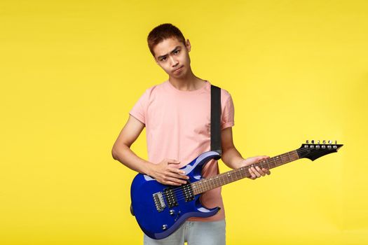 Lifestyle, leisure and youth concept. Gloomy asian guy playing in band, look sad and uneasy, have bad day, holding electric guitar, trying learn new instrument, stand yellow background.