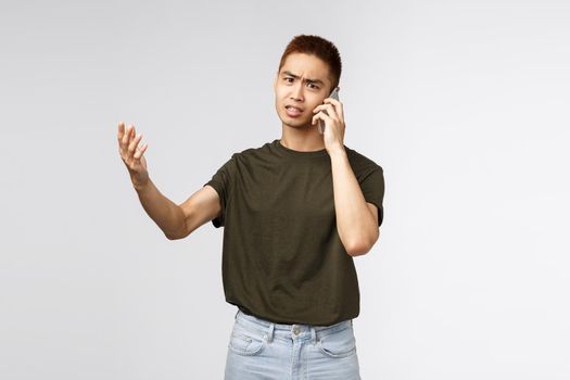 Technology, online lifestyle and communication concept. Portrait of concerned, complaining asian man shaking hands in dismay, arguing via phone call, react displeased to bad news. Copy space