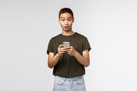 Technology, online lifestyle and communication concept. Portrait of surprised and confused asian guy asking question as receive strange message, holding mobile phone and look indecisive.