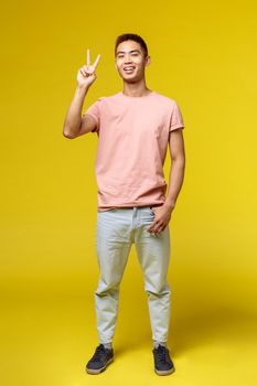 Lifestyle, education and people concept. Vertical portrait of stylish young asian guy in summer outfit, show peace sign and smiling, greeting people at party, standing enthusiastic yellow background.