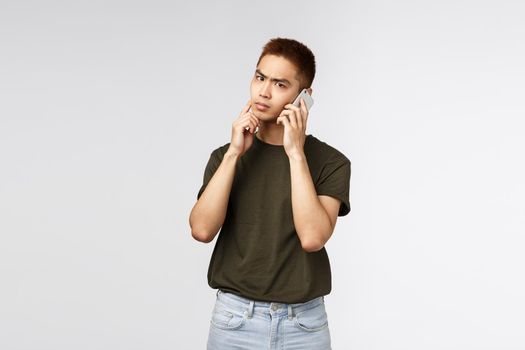 Technology, online lifestyle and communication concept. Thoughtful serious asian man talking on phone, making important decision, touch chin and frowning, thinking as make choice during conversation.