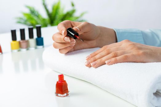 Woman giving herself elegant manicure at home. Closeup of beautiful female hand applying red nail polish. Colorful nail polish bottles on table. Nail care and beautician procedure. Stylish nail art