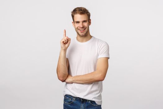 Guy made up excellent idea. Attractive smiling blond man in white t-shirt, bristle, raise index finger, pointing up or have suggestion, express own opinion, make eureka gesture, white background.