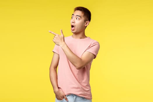 People, different emotions and lifestyle concept. Portrait of shocked asian guy in pink t-shirt, gasping, drop jaw from amazement, stare startled and pointing finger left, standing yellow background.