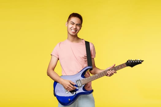 Lifestyle, leisure and youth concept. Lets jam. Carefree smiling asian guy playing in band, holding blue electric guitar, feel rock-n-roll start on stage, standing upbeat yellow background.