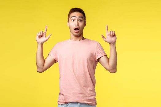 Lifestyle, people and summer vacation concept. Surprised, impressed asian male in pink t-shirt, pointing fingers up, open mouth astonished and amazed, showing new advertisement, cool product.