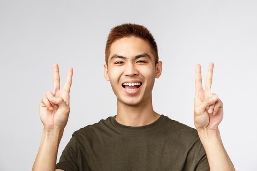 People, different expressions and lifestyle concept. Close-up portrait of joyful handsome young male student, japanese guy feel upbeat, getting ready for vacation, show peace signs and smile happy.