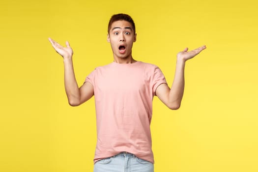 Vacation, summer and people concept. Portrait of surprised handsome, startled asian man gasping, raise hands sideways in amazement and awe, react to wonderful astonishing news, yellow background.