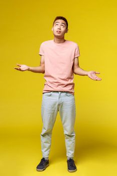 Lifestyle, education and people concept. Vertical portrait of confused shrugging asian man, looking up at sky, asking God why, dont know and cant tell, stand yellow background.