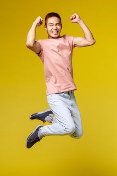 Summer vacation, education and lifestyle concept. Vertical shot of happy athletic asian guy, flex biceps and smiling upbeat, jumping from joy, triumphing, celebrating victory, yellow background.