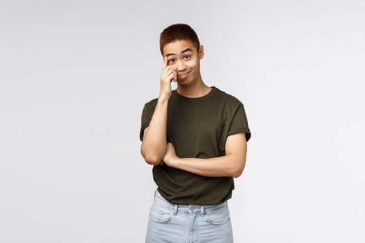 Portrait of bothered and annoyed asian guy hear nonsense, smirk and stare with disbelief, scratching forehead and look camera ignorant, standing grey background distressed and tensed.