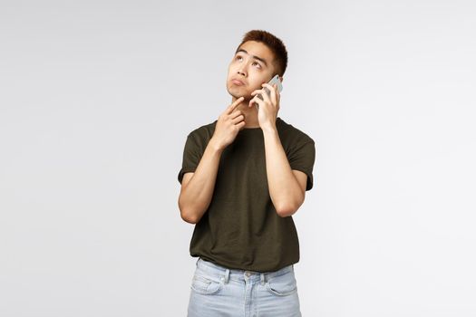 Technology, online lifestyle and communication concept. Portrait of unsure thoughtful asian man making decision, order food delivery via phone call, touch chin, ponder look up, talk on smartphone.