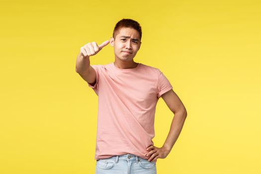 Portrait of skeptical displeased asian man in pink t-shirt, judging, express own opinion to bad thing, rate product is average, thumb-down and grimacing disappointed, yellow background.