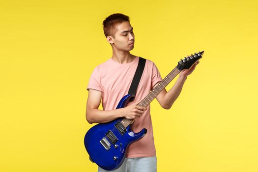 Lifestyle, leisure and youth concept. Portrait of good-looking asian guy playing in band, tune electric guitar before coming on stage and perform song, stand yellow background.