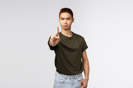 Portrait of strict and serious, angry asian man shaking finger, scolding person for doing bad think, disobey rules, frowning disappointed, prohibit or disapprove behaviour, grey background.