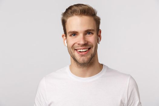 Handsome dreamy and excited blond caucasian guy with sexy bristle, wear wireless headphones and smiling, speaking, talking on phone using earphones, listen music earbuds, enjoy podcast.