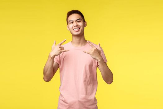 Portrait of cool and proud good-looking asian guy with big ego, show rock-n-roll or heavy metal sign and smiling, enjoying awesome concert, having fun on music festival, yellow background.