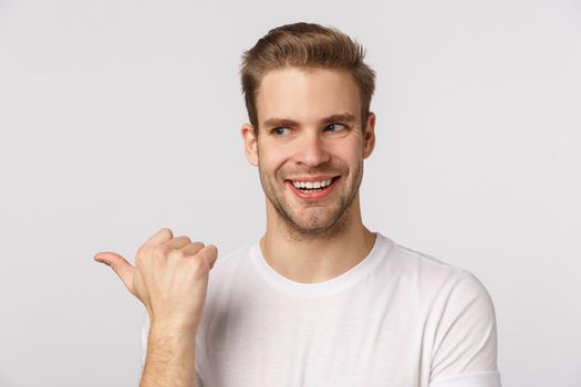 Intrigued and sly good-looking blond bearded guy with bristle in white t-shirt, peeking and pointing left with excited, thrilled smile, standing enthusiastic, check out something desirable.
