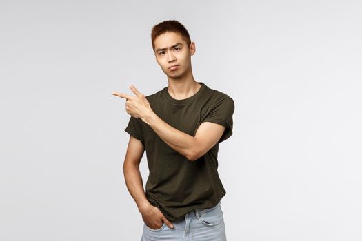 Portrait of skeptical, gloomy young asian male student hate self-distancing from friends and college pals, pointing finger left and grimacing camera, pouting upset, feel envy or regret.