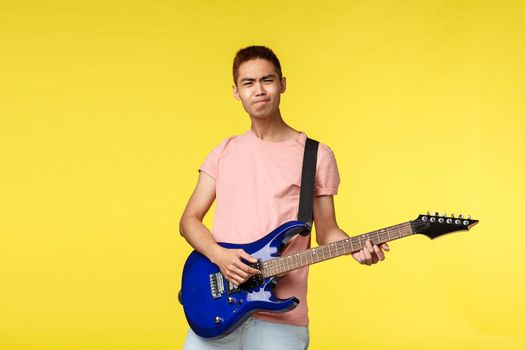 Lifestyle, leisure and youth concept. Enthusiastic carefree asian man in pink t-shirt playing on electric guitar over yellow background, enjoying performing with band, learn new instruments.