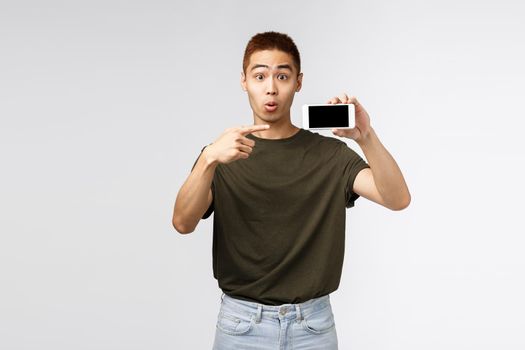 Technology, online lifestyle and communication concept. Portrait of curious and interested asian man asking question about app or internet store, pointing finger at smartphone, grey background.