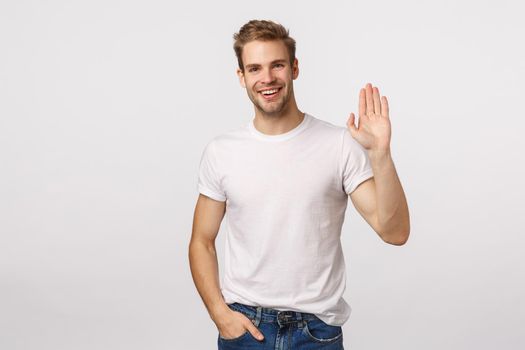 Friendly, handsome macho man in white t-shirt, blond hair and bristle, raise one hand, waving and smiling happy, meeting friend in pub, say hello or hi, greeting, standing white background.
